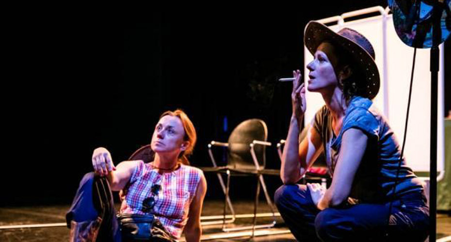 Drama students performing on stage, girl crouching down smoking and wearing a cowboy hat. Other girl looking into the distance.