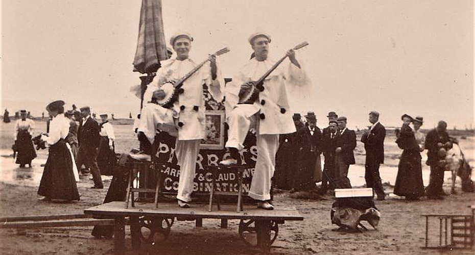 Black and white photograph - two men standing on a platform playing banjos on the beach, with a crowd behind watching.